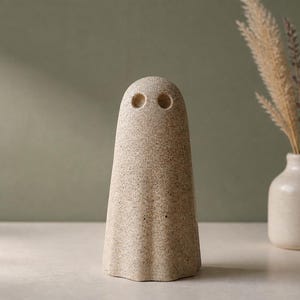 May include: A beige, speckled, ghost-shaped figurine with a smooth, rounded top and two circular indentations for eyes. The object has a flared base and a textured, stone-like appearance, standing upright as a singular decorative piece.