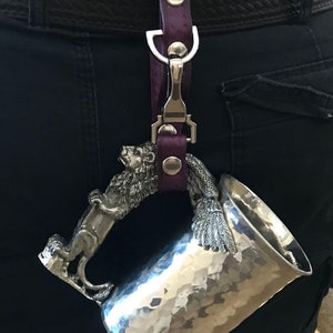 May include: A silver metal mug with a lion and bird handle. The mug is attached to a purple leather strap with a silver clip.