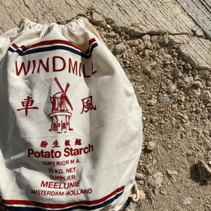 May include: A white canvas bag with red and blue stripes. The bag has a windmill design with the text "WINDMILL" in red. The bag also has the text "Potato Starch" in red, along with the words "SUPERIOR M A." and "25 KG. NET". The bag also has the text "SUPPLIER: MEELUNIE AMSTERDAM-HOLLAND" in red.