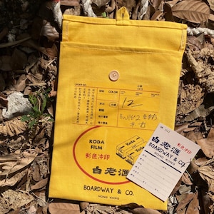 May include: A yellow canvas bag with a flap and a button closure. The bag has a label that says "Koda Film 彩色冲印 白老酒 BOARDWAY & CO. HONG KONG". There is a smaller white label with the text "白老酒 彩色冲印 BOARDWAY & CO. HONG KONG" and a handwritten note that says "BW×2 各打 共打 12".