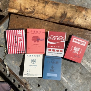 Può includere: Cinque quaderni in stile vintage con disegni rossi, bianchi e blu. I quaderni presentano testo in cinese e inglese, tra cui "Hong Kong Bank", "Cold Coke", "Ristorante dell'aeroporto", "Manuale dello studente" e "Gas di petrolio".
