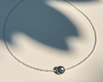 Op de afbeelding: Een delicate witte kralenketting met een enkele grote, donkergrijze parel hanger. De ketting ligt op een wit oppervlak met een schaduw.