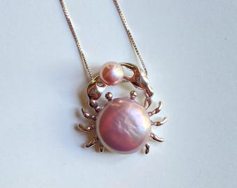 May include: A silver crab pendant necklace with a pink pearl in the center and a smaller pink pearl on the claw. The necklace is on a silver chain.