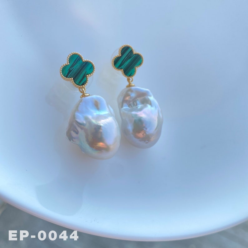 May include: A pair of white baroque pearl drop earrings with green malachite clover charms. The earrings are on a white surface.