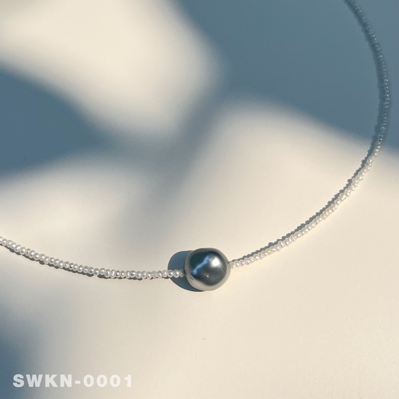 May include: A delicate necklace with a single, large, gray pearl pendant. The necklace is made of small, white beads.