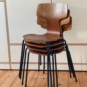 Könnte beinhalten: Vier Holzstühle mit schwarzen Metallbeinen, die übereinander gestapelt sind. Die Stühle sind aus hellbraunem Holz gefertigt und haben ein schlichtes, modernes Design.