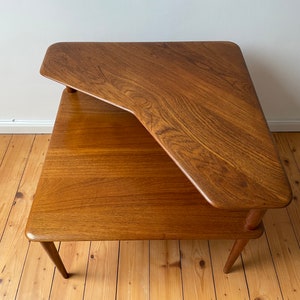 Könnte beinhalten: Ein zweistufiger Ecktisch aus massivem Holz mit natürlicher Oberfläche. Die obere Ebene ist dreieckig, die untere rechteckig. Der Tisch hat ein schlichtes, modernes Design aus der Mitte des Jahrhunderts.