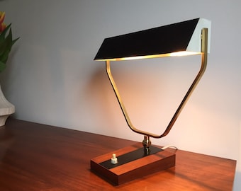 Könnte beinhalten: Eine Schreibtischlampe im Stil der Jahrhundertmitte mit einem schwarz-weißen Schirm und einem goldenen Metallrahmen. Die Lampe hat einen Holzsockel mit einem schwarzen Schalter.