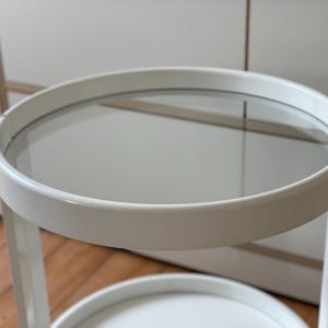 May include: A white, two-tiered side table with a circular design. The top tier features a clear glass surface, and the lower tier is solid white. The table is supported by white legs and is suitable for various home decor styles.