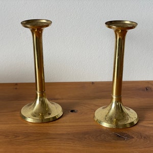 Op de afbeelding: Twee gouden metalen kaarsenstandaards met een eenvoudig ontwerp. De standaards hebben een ronde basis en een hoge, cilindrische steel.