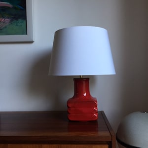 May include: A red ceramic table lamp with a white lampshade. The lamp is on a wooden table with two wooden candle holders.
