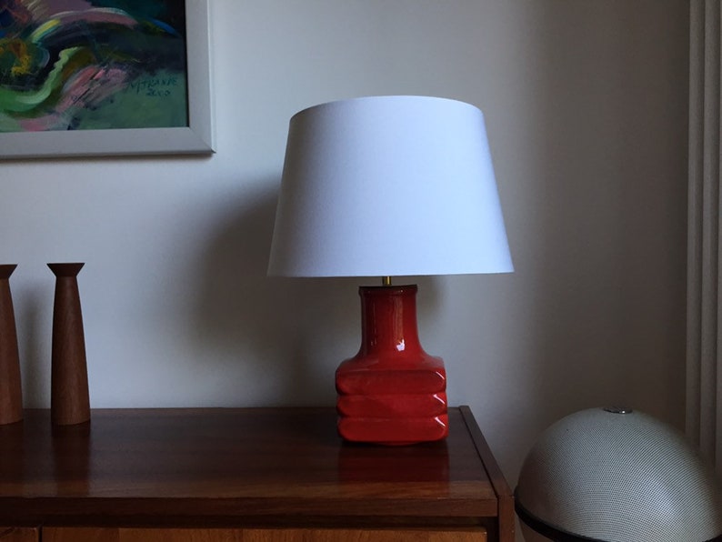 May include: A red ceramic table lamp with a white lampshade. The lamp is on a wooden table with two wooden candle holders.