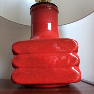 May include: A red ceramic table lamp with a white lampshade. The base of the lamp is shaped like a rectangle with rounded edges and has a textured surface.