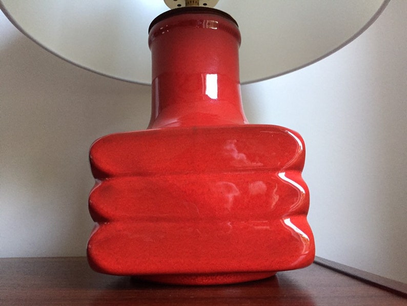 May include: A red ceramic table lamp with a white lampshade. The base of the lamp is shaped like a rectangle with rounded edges and has a textured surface.