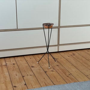 Op de afbeelding: Een koperkleurige kom staat op een zwarte metalen standaard met drie poten. De standaard staat op een houten vloer.