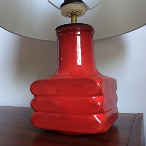 May include: A red ceramic table lamp with a white lampshade. The lamp base has a unique, layered design.