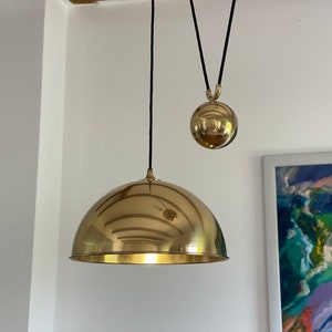 Peut inclure: Un luminaire suspendu en or avec un grand abat-jour rond en forme de dôme et un petit poids rond en forme de boule suspendu à un cordon noir.