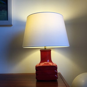 May include: A red ceramic table lamp with a white lampshade. The lamp is on a wooden surface.