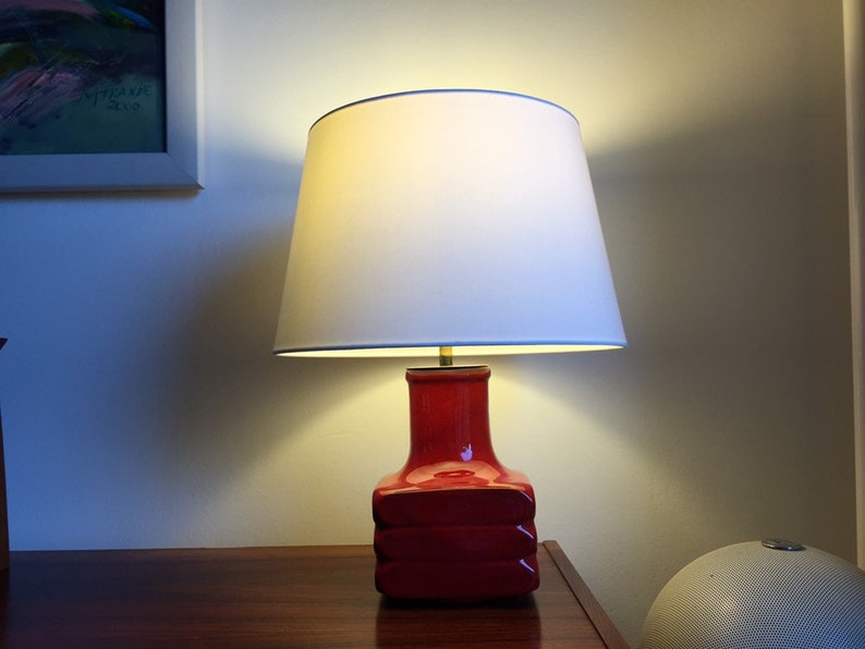 May include: A red ceramic table lamp with a white lampshade. The lamp is on a wooden surface.