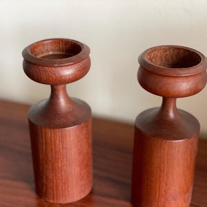 Op de afbeelding: Twee roodbruine houten kandelaars. Elke kandelaar heeft een cilindrische basis, een slanke steel en een cirkelvormige bovenkant voor een kaars. De houtnerf is zichtbaar.