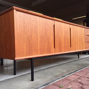 May include: A mid-century modern teak sideboard with black metal legs. The sideboard has two doors and three drawers. The wood grain is visible on the surface of the sideboard.