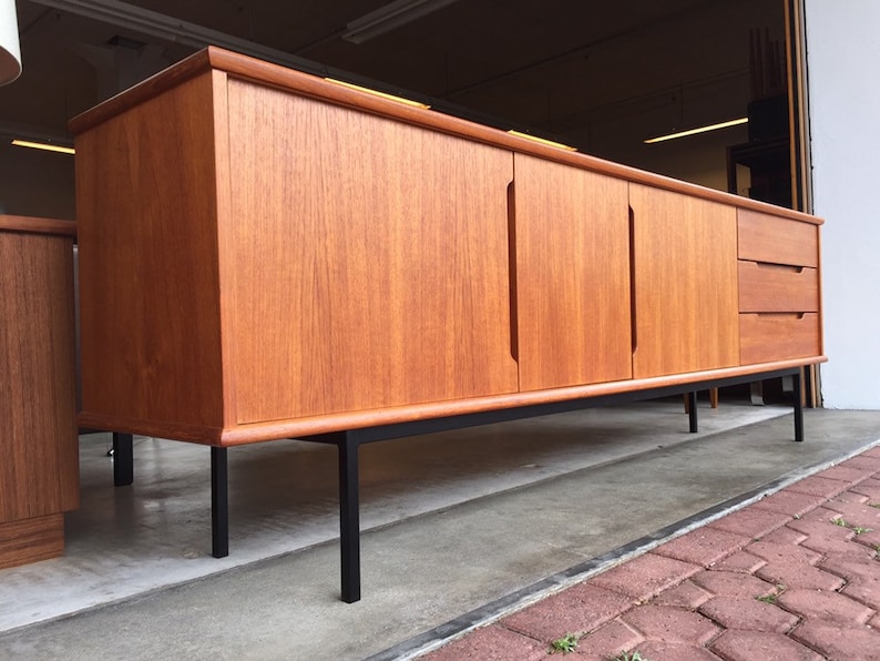 May include: A mid-century modern teak sideboard with black metal legs. The sideboard has two doors and three drawers. The wood grain is visible on the surface of the sideboard.