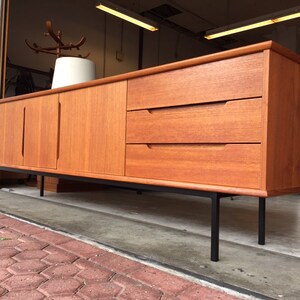 May include: A mid-century modern teak sideboard with three drawers and two doors. The sideboard has black metal legs and a light wood finish.