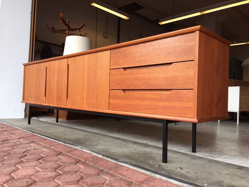May include: A mid-century modern teak sideboard with three drawers and two doors. The sideboard has black metal legs and a light wood finish.