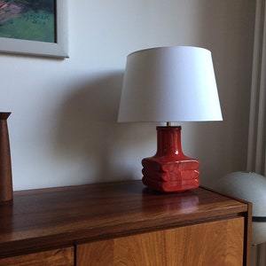 May include: A red ceramic table lamp with a white lampshade. The lamp is sitting on a wooden table.