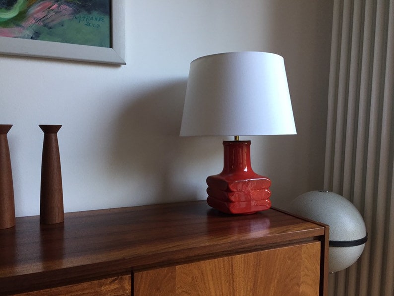 May include: A red ceramic table lamp with a white lampshade. The lamp is sitting on a wooden table.