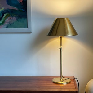 以下が含まれることがあります： 丸い金色のシェードが付いた金色のテーブルランプ。ランプは木製のテーブルの上にあり、背景は白い壁です。