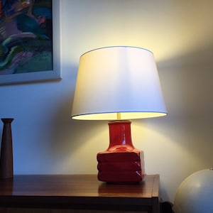 May include: A red ceramic table lamp with a white lampshade. The lamp is on a wooden table with two wooden candle holders.