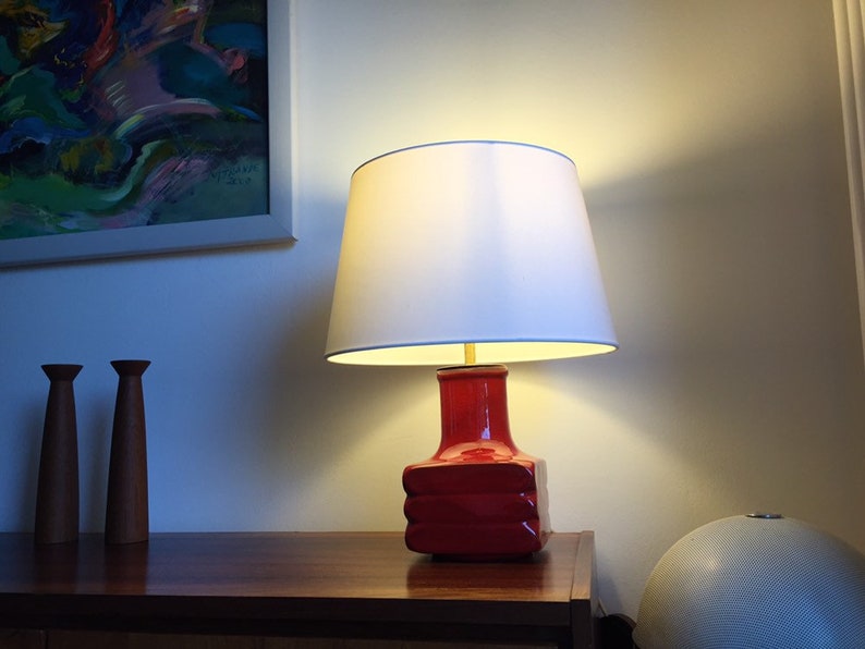 May include: A red ceramic table lamp with a white lampshade. The lamp is on a wooden table with two wooden candle holders.