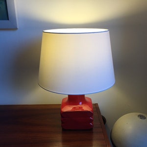 May include: A red ceramic table lamp with a white fabric lampshade. The lamp is switched on and illuminating the room.