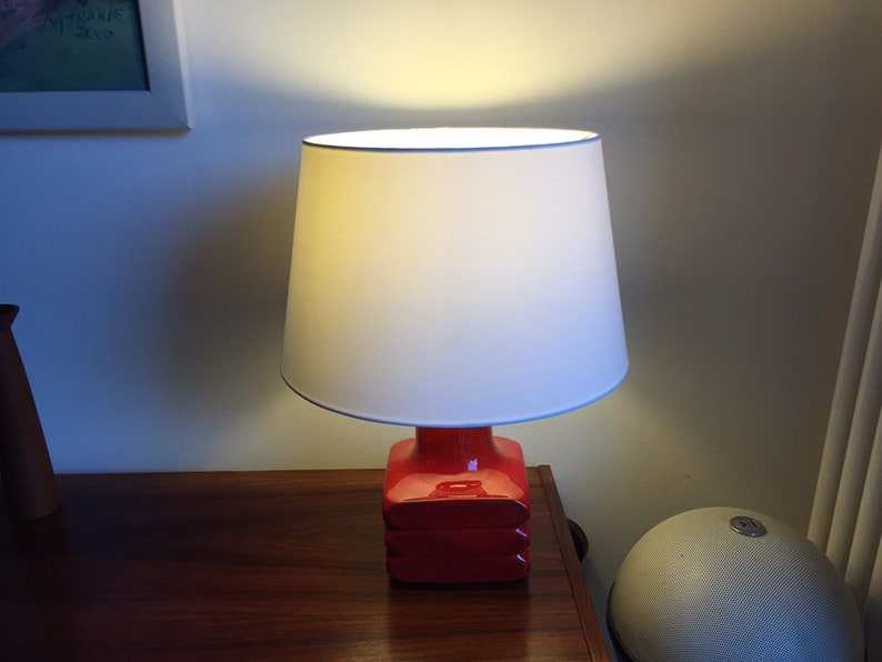 May include: A red ceramic table lamp with a white fabric lampshade. The lamp is switched on and illuminating the room.