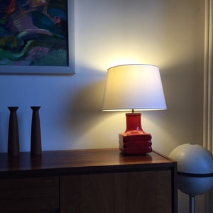 May include: A red ceramic table lamp with a white lampshade. The lamp is on a wooden table with two wooden candle holders.