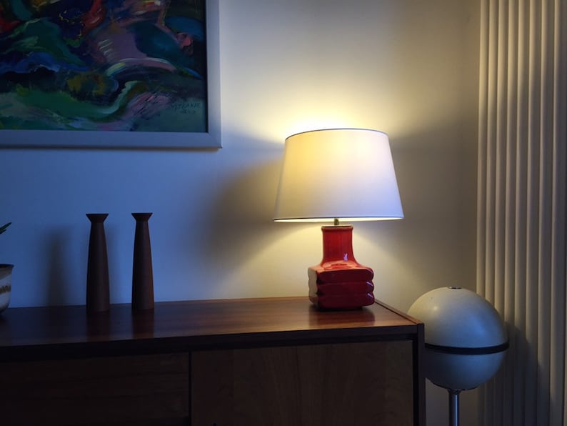 May include: A red ceramic table lamp with a white lampshade. The lamp is on a wooden table with two wooden candle holders.