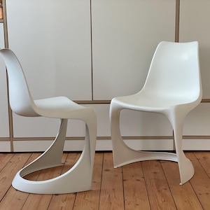 Könnte beinhalten: Zwei weiße Plastikstühle mit einem einzigartigen, geschwungenen Design. Die Stühle sind aus glattem, weißem Kunststoff gefertigt und haben eine moderne, minimalistische Ästhetik.