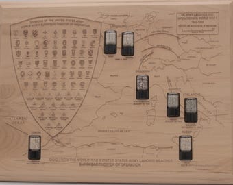May include: A light-coloured wooden plaque featuring an engraved map of the European theatre of operations during World War II. It includes a shield with military division insignias and eight small black containers filled with sand from landing beaches.