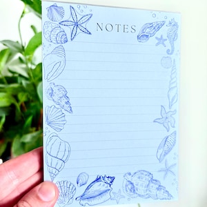 May include: A pale blue notepad featuring a border of blue seashell illustrations and the word "NOTES". The notepad has lined pages. A hand is holding the notepad.