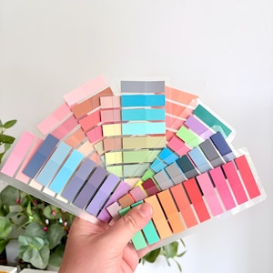 May include: A collection of translucent adhesive page markers arranged in a fan shape. The rectangular strips come in a wide variety of colors including pink, blue, green, orange, purple, gray, and red, all mounted on clear plastic backing sheets.