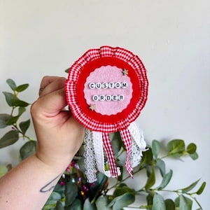 May include: A circular rosette badge featuring a red and white gingham pleated border, a scalloped pink felt center, and the text CUSTOM ORDER spelled out in black and white plastic letter beads, accented with two small gold star embellishments.
