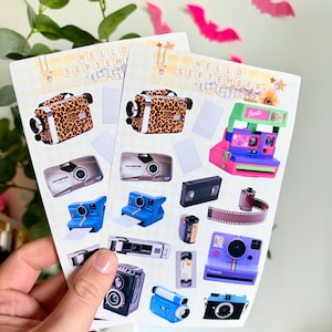 May include: Two sheets of stickers with camera designs, including a leopard print camcorder, a pink and purple Polaroid, and a blue instant camera. The stickers have the text "HELLO SEPTEMBER". Pink bat cutouts are in the background.