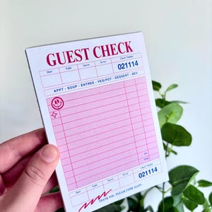 May include: A pink and white guest check notepad with the words "GUEST CHECK" in large, red letters. The notepad has sections for date, table, guests, and server, with a check number of 021114. The bottom reads "THANK YOU, PLEASE COME AGAIN."