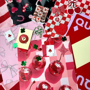 May include: A selection of decorative paper and embellishments in red, pink, and black. Designs include cherries, bows, playing cards, disco balls, and lucky charms. The items are arranged on a red and white striped surface.