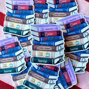 Op de afbeelding: Stickers met afbeeldingen van stapels boeken met titels als "romantasy", "smut", "mystery", "cowboy romance", "cozy reads" en "horror". De boeken hebben verschillende kleuren en decoratieve elementen, tegen een rood-wit gestreepte achtergrond.