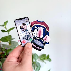 May include: A collection of stickers featuring a smartphone with a video call, a hockey jersey with the name "Graham" and the number 44, and a hockey puck with pink bunny ears. The phone screen displays a couple on ice skates.