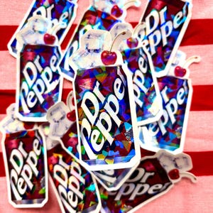 May include: A collection of Dr Pepper can stickers with a cherry on top. The stickers have a holographic, geometric design and the Dr Pepper logo in white. The background is a red and white striped fabric.