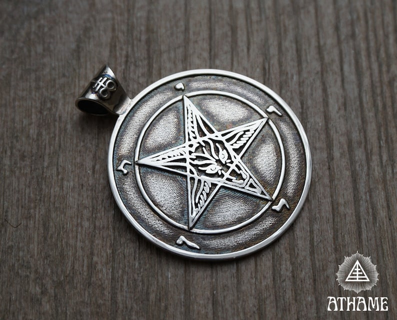May include: Silver pendant featuring a pentagram with a goat's head inside, surrounded by a circle of symbols. The pendant is on a wooden surface.