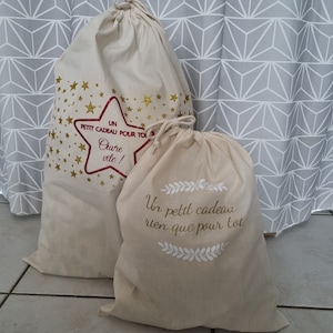Puede incluir: Dos bolsas de regalo de tela beige con letras doradas. La bolsa más grande tiene estrellas doradas y el texto "UN PETIT CADEAU POUR TOI Oure vite!" La bolsa más pequeña tiene un diseño de corona blanca y el texto "Un petit cadeau rien que pour toi".